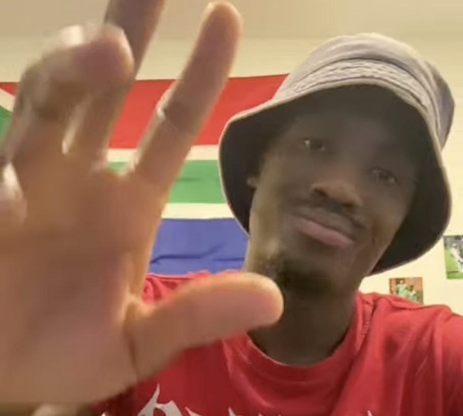 Smiling student-athlete wearing a bucket hat waves during testimonial with South African flag in the background.